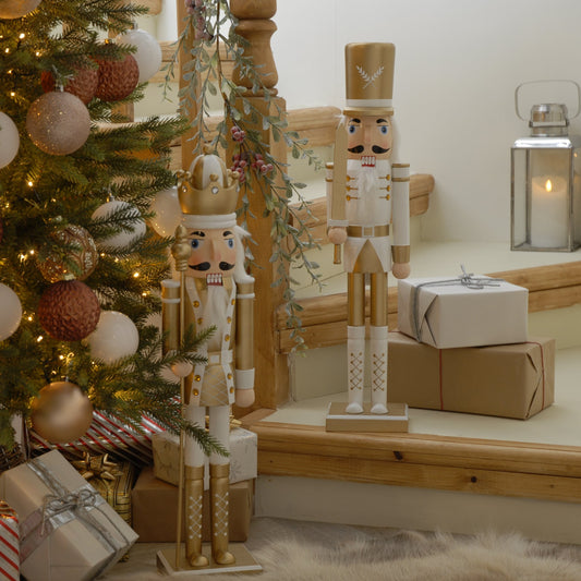 Two white and gold nutcrackers standing on a wooden surface with a Christmas tree and gifts in the background.