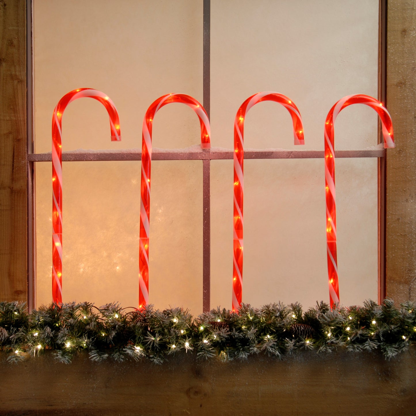 A set of four red LED lit candy cane stake lights placed in a garden, visible through a window, with a transformer included for indoor use.