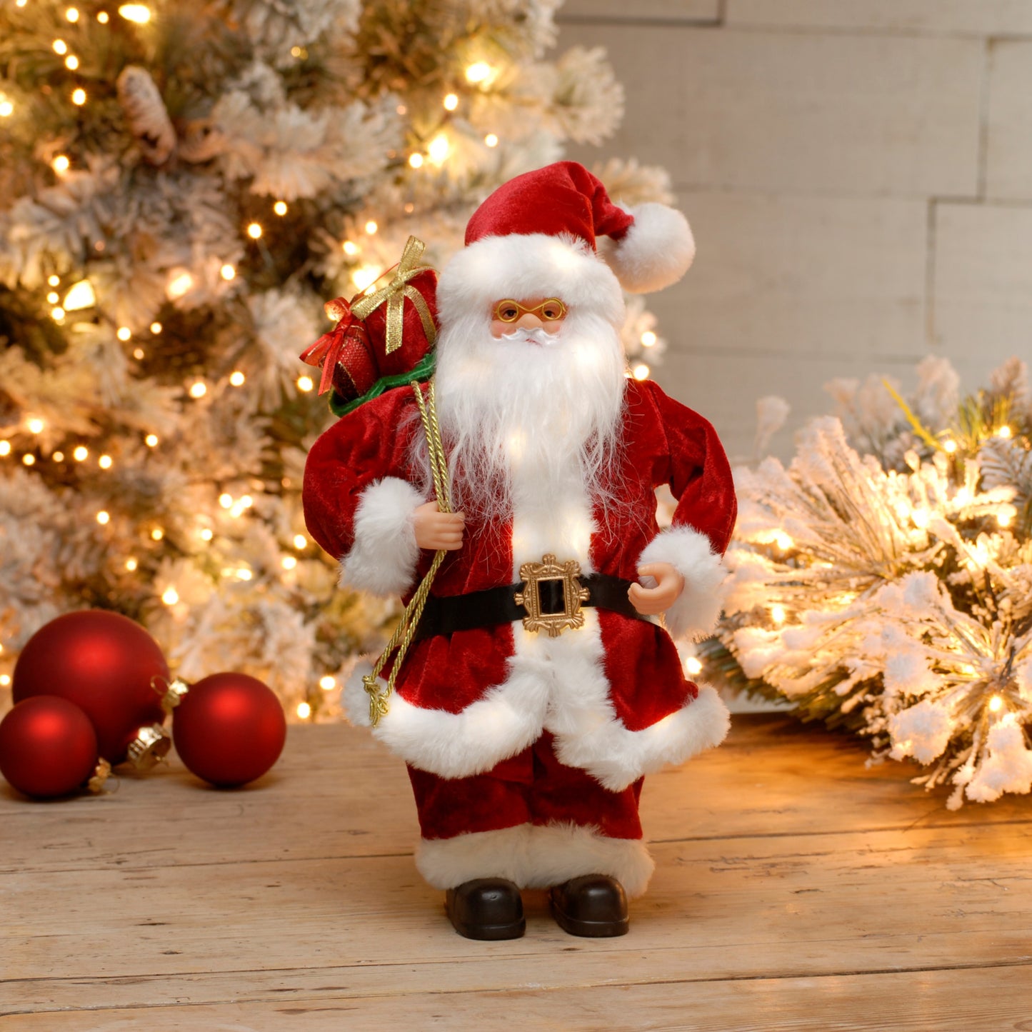 A battery-operated, lighted classic Santa figurine standing on a wooden surface with Christmas decorations in the background.
