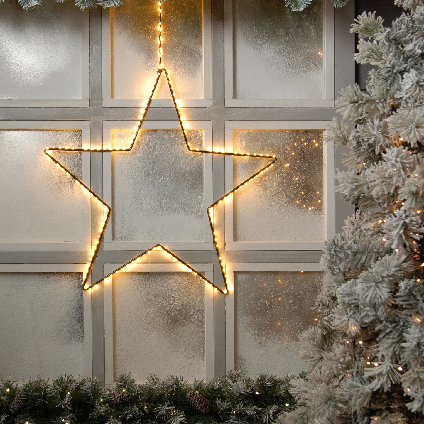 A 58cm LED lit black star-shaped decoration with warm white lights, displayed on a window sill, surrounded by a frosty scene with Christmas trees in the background.