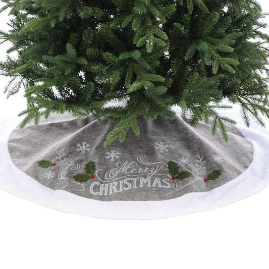 A grey Christmas tree skirt with 'Merry Christmas' text and holly and snowflake design, placed under a green Christmas tree.