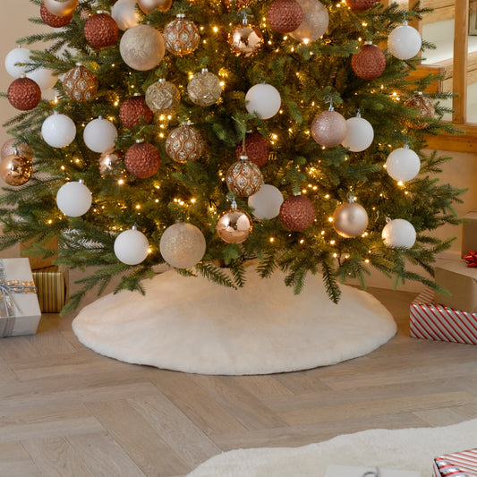 A white faux fur Christmas tree skirt placed under a decorated Christmas tree, with gifts around it on a wooden floor.