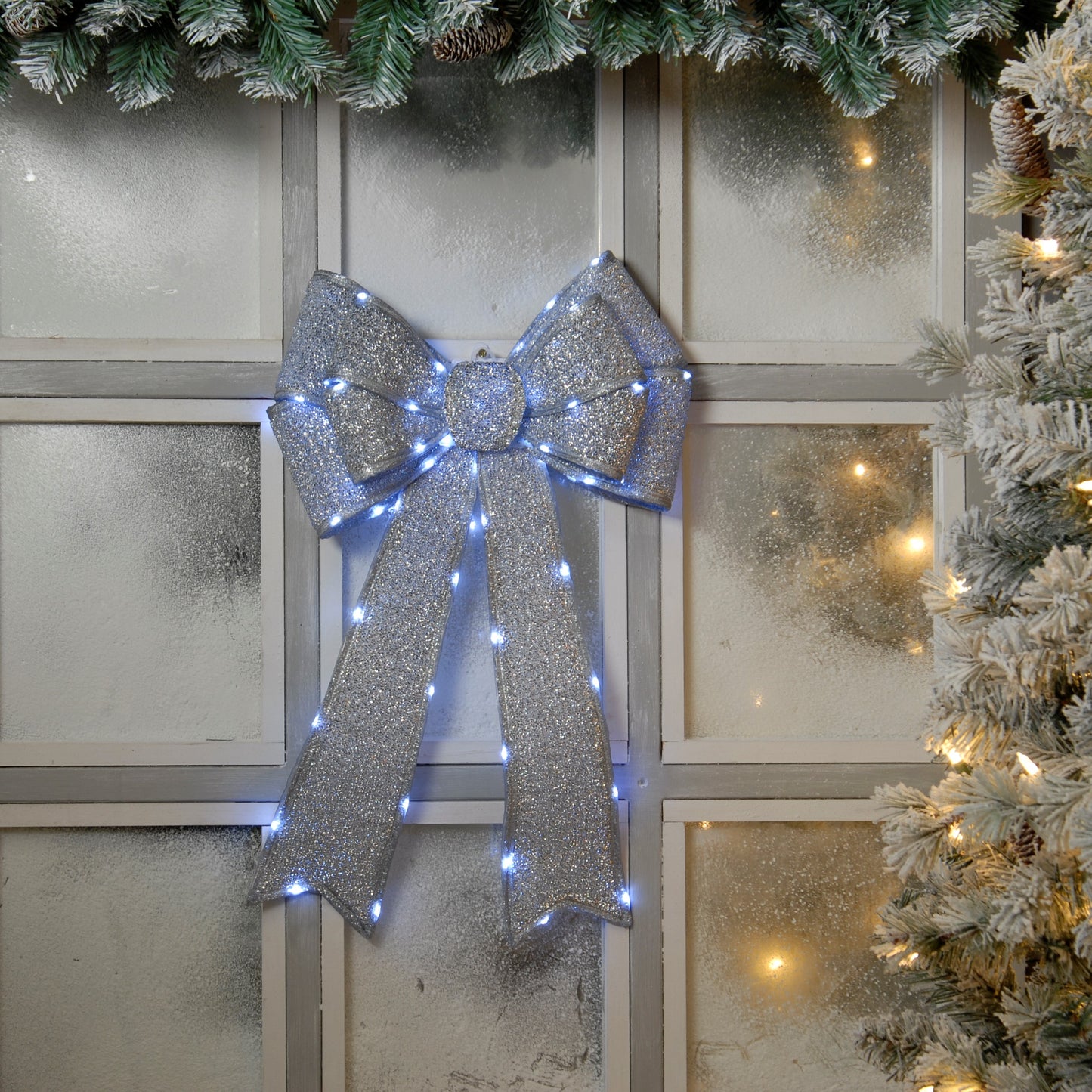 A silver fabric Christmas bow with battery-operated white LED lights, displayed on a window with a festive wreath in the background.
