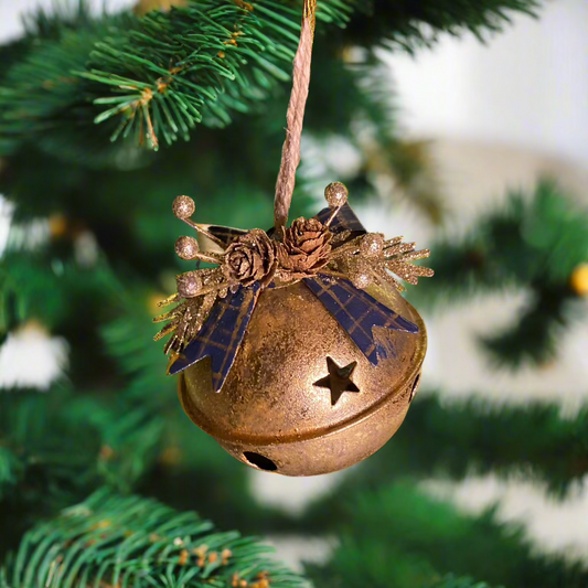 A gold-colored metal sleigh bell tree decoration with a rustic style, aged effect, and blue metal ribbon, hanging from a green pine tree.