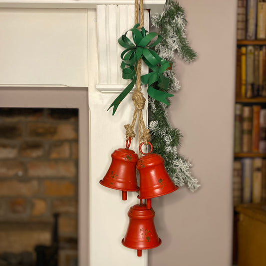 Three red Christmas bells with green metal ribbons and a rustic jute rope, hanging from a brick wall with a decorative fireplace and Christmas tree in the background.