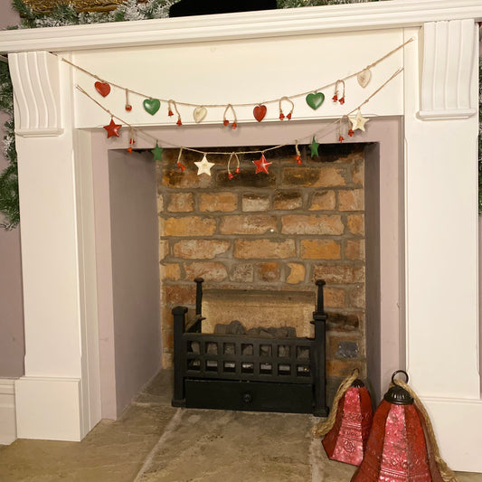 A garland of red, white, and green hearts and stars hanging over a fireplace, with Christmas tree decorations in the background.