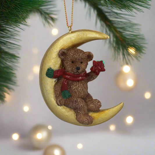 Decorative ornament of a teddy bear sitting on a crescent moon, holding a red star, on a white background.