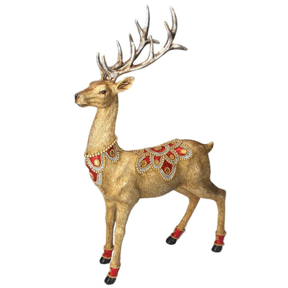 A gold-colored Christmas stag ornament adorned with red jewels on its saddle, collar, and anklets, placed against a backdrop of Christmas lights and a pine tree.