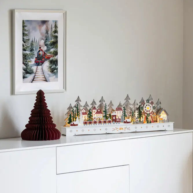 A Christmas decoration featuring a wooden structure with LED lights resembling a village, placed on a white surface, with a framed picture of a winter scene hanging above it.