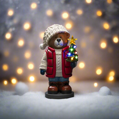 Bear figurine in winter clothing holding a small Christmas tree with lights against a blurred light background.