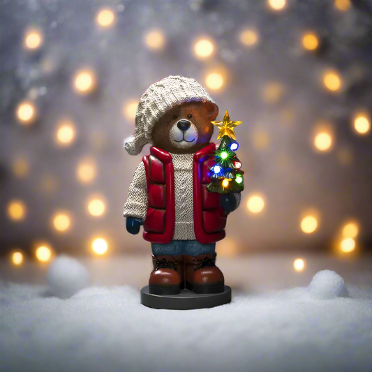 Bear figurine in winter clothing holding a small Christmas tree with lights against a blurred light background.