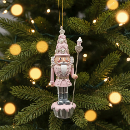 A pink cupcake nutcracker hanging from a string, with a Christmas tree and lights in the background.