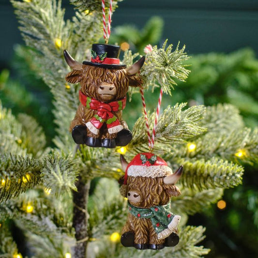 Two resin Christmas ornaments featuring Highland cows with festive scarves and hats, hanging on a Christmas tree.