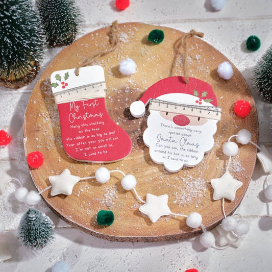 Two wooden Christmas keepsake hangers, one designed with a Santa Claus face and the other with a Christmas message, decorated with white and red pom-poms, presented on a circular wooden board with festive decorations around.