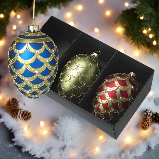 A set of three ornate glass Christmas tree baubles in blue, green, and red, with gold designs, displayed in a black box.