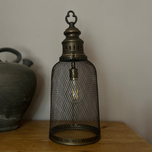 A bronze mesh metal lantern with a glass cover and a visible LED light source, placed on a wooden surface.