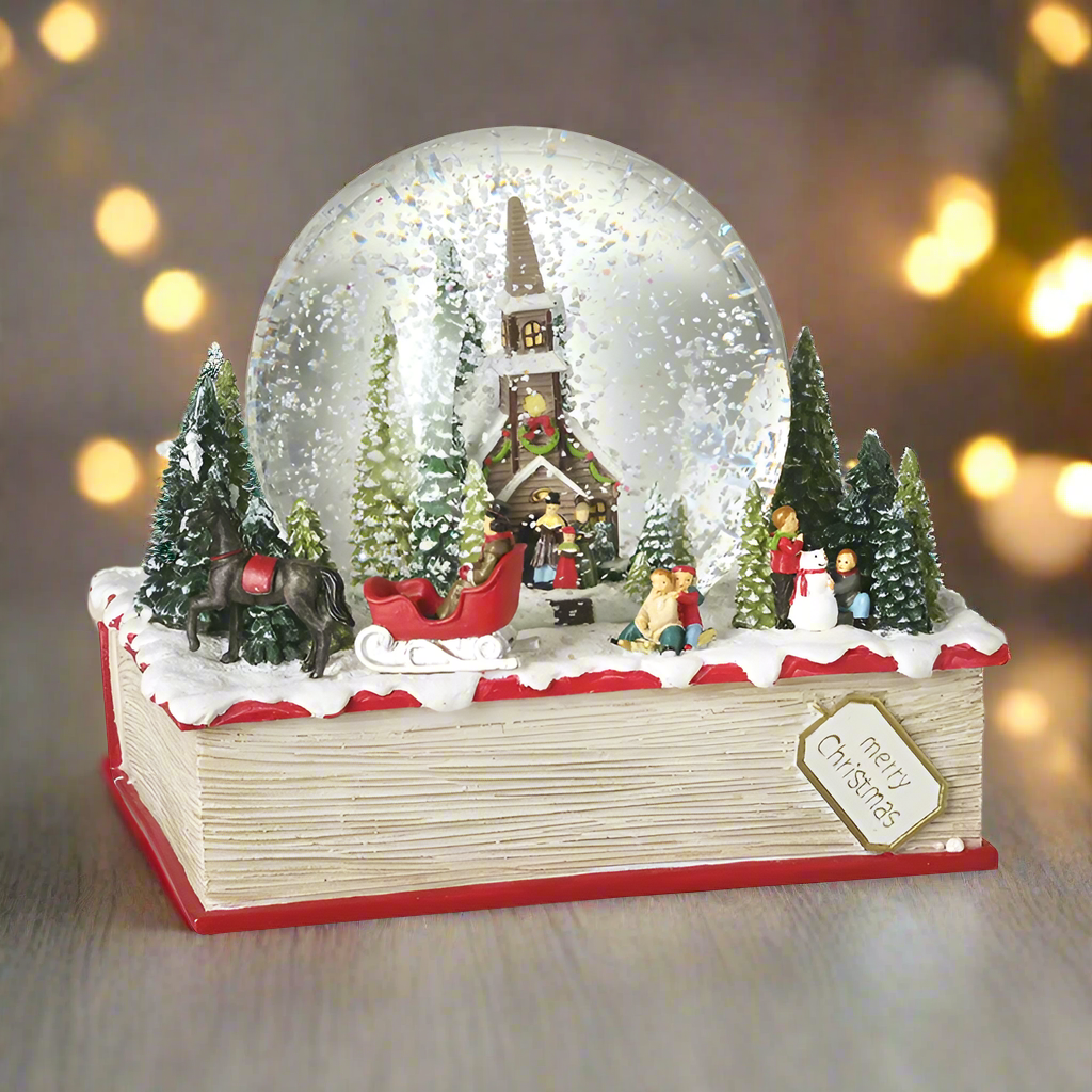 A resin snow globe featuring a Christmas church scene with trees, inside the globe snow is visible. The base of the globe is designed to look like a red and white book with holiday decorations on top.