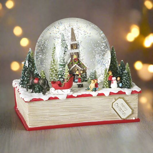 A resin snow globe featuring a Christmas church scene with trees, inside the globe snow is visible. The base of the globe is designed to look like a red and white book with holiday decorations on top.
