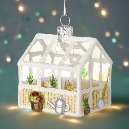 A glass ornament in the shape of a greenhouse with plants and tools inside, hanging from a silver loop.