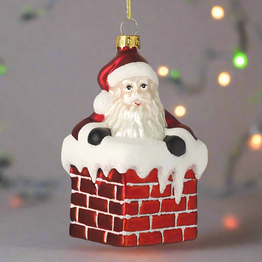 A glass decoration featuring Santa Claus with a red suit and white beard, peeking out from a red chimney with white snow on it, designed for hanging on a Christmas tree.