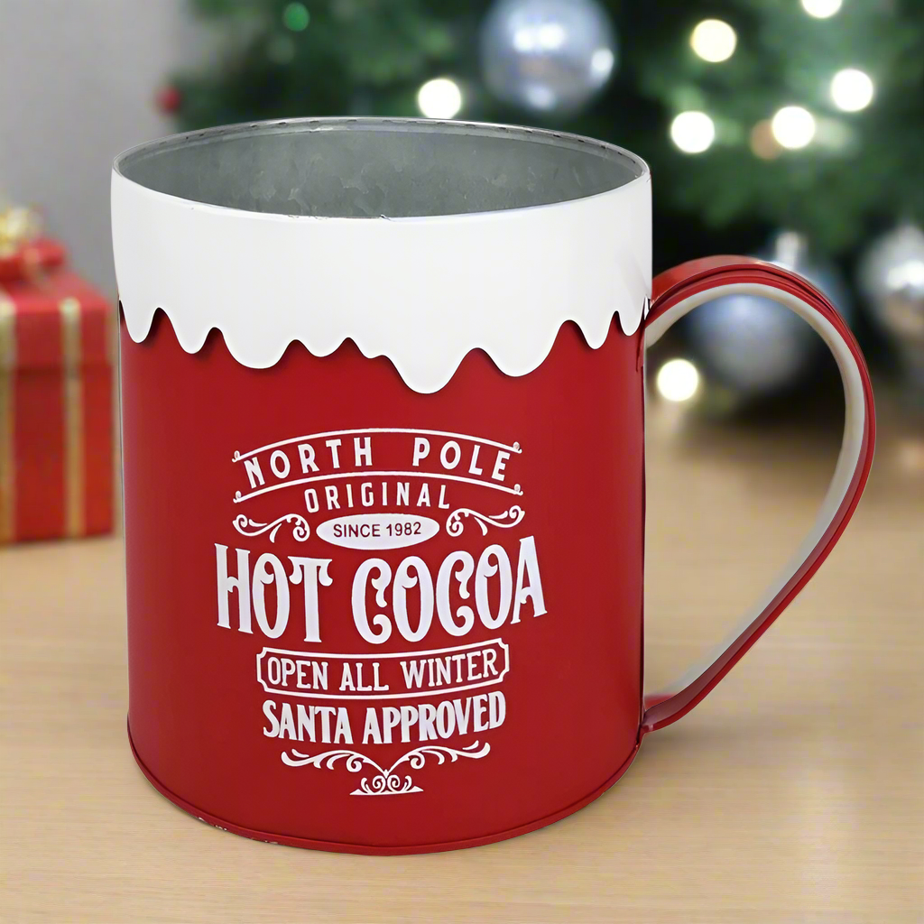 A large display red and white metal mug with 'North Pole Hot Cocoa' text and a snowflake design on the rim.
