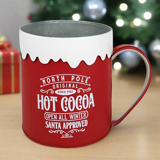 A large display red and white metal mug with 'North Pole Hot Cocoa' text and a snowflake design on the rim.