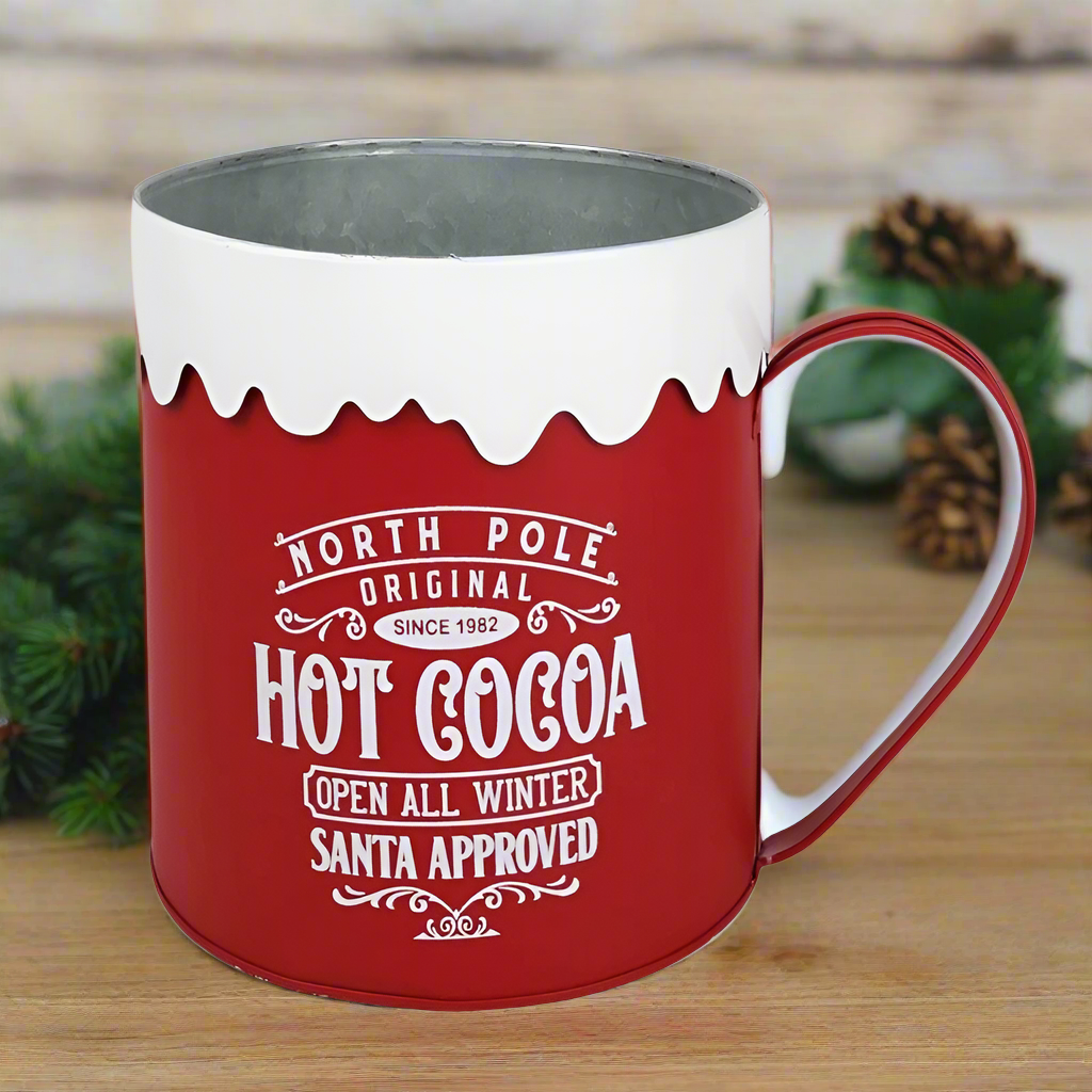 A red and white metal mug with 'North Pole Hot Cocoa' text and a snowflake design on the rim.