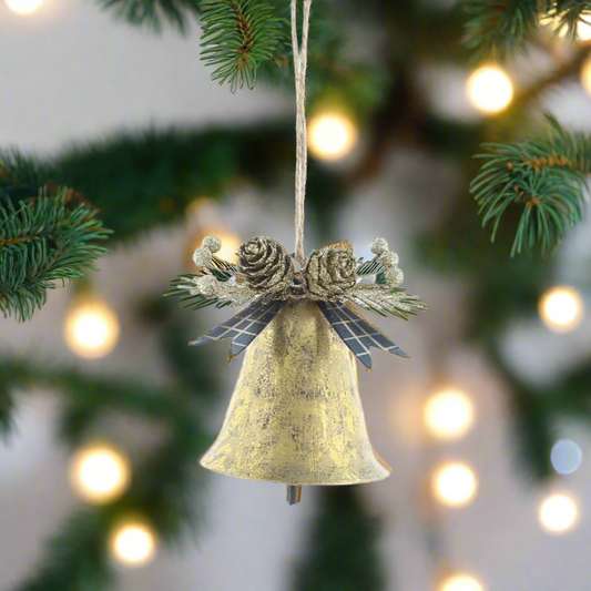 A gold-colored metal bell with a vintage effect, hanging from a string, decorated with aged foliage and a seasonal bow, displayed against a blurred Christmas tree background.