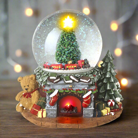A musical snowglobe featuring a Christmas fireplace scene with a tree inside the globe, surrounded by holiday decorations and a teddy bear.